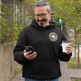 a man standing on the sidewalk looking at his phone and wearing a black hoodie with the Certified Caffeiniac design on the front left chest