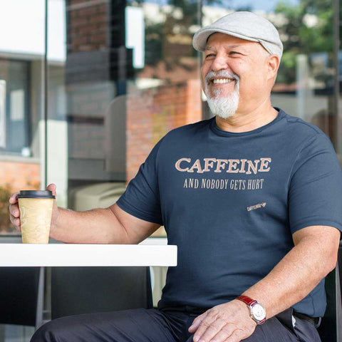 Image of Smiling man sitting down in a coffee shop wearing a navy blue Caffeiniac t-shirt with the design CAFFEINE and nobody gets hurt 