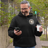 a man standing on the sidewalk looking at his phone and wearing a black hoodie with the Certified Caffeiniac design on the front left chest