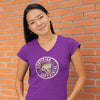 smiling woman standing against a brick wall wearing a purple v-neck shirt featuring the Certified Caffeiniac design on the front