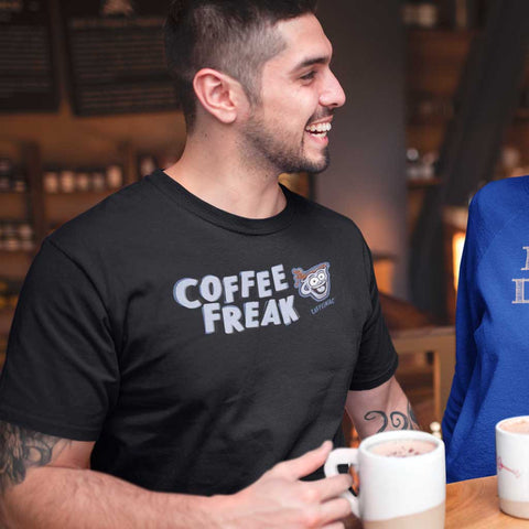 Image of smiling man having a cup of coffee in a coffee shop wearing a black Caffeiniac t-shirt featuring the Coffee Freak design on the frontt