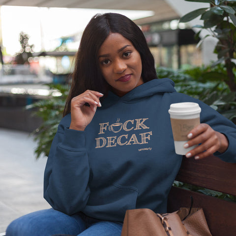 Image of woman on a bench wearing a navy blue hoodies sweatshirt holding a cup of coffee. The front of the sweatshirt features the Cafffeiniac design F_CK DECAF