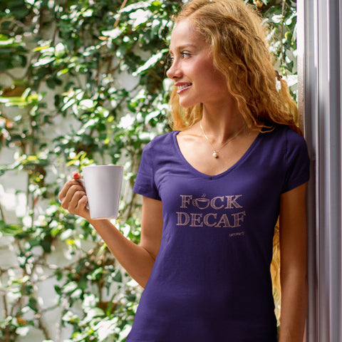 Image of woman standing in a patio enjoying a cup of coffee, wearing a purple v-neck shirt with the Caffeiniac design F_CK DECAG in tan ink