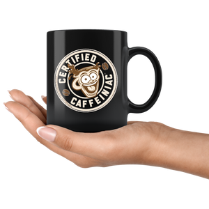 a black coffee mug featuring the Certified Caffeiniac design in tan and brown printed on the front and back resting on a woman's palm